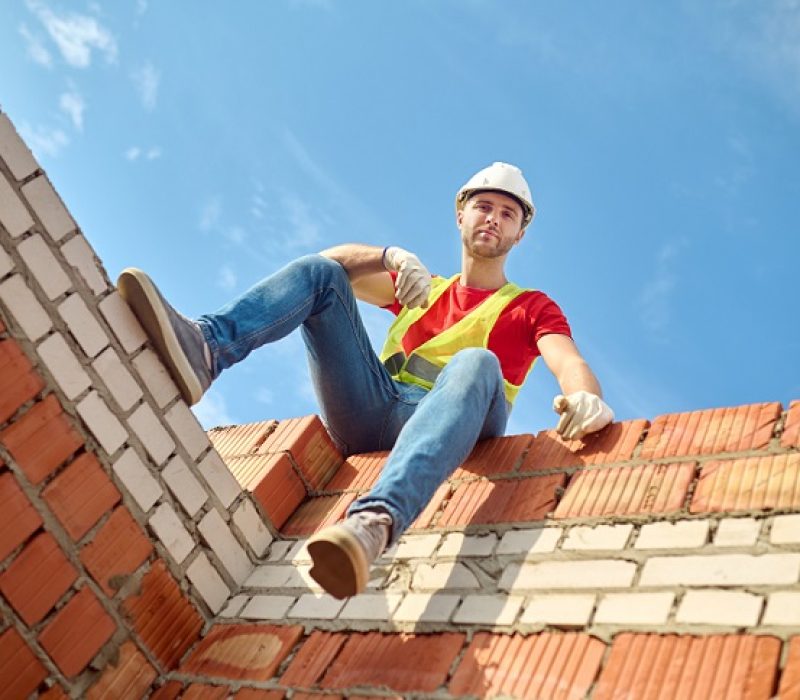 Short break. Bottom view of man in protective helmet and gloves sitting on brick wall looking at camera on sunny day at construction site